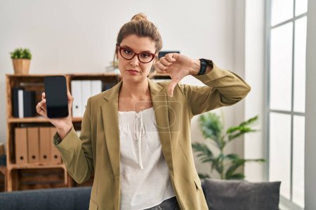 Foto de Young woman working at consultation office holding smartphone with angry face, negative sign showing dislike with thumbs down, rejection concept - Imagen libre de derechos