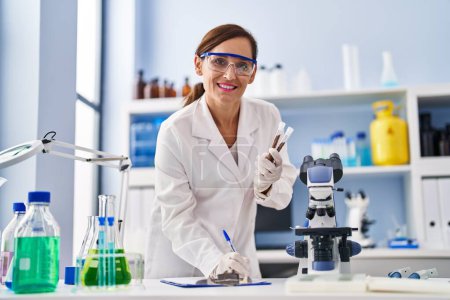 Téléchargez les photos : Middle age woman wearing scientist uniform writing on clipboard holding test tubes at laboratory - en image libre de droit