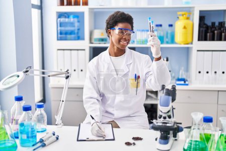 Photo for African american woman wearing scientist uniform write on clipboard holding test tubes at laboratory - Royalty Free Image