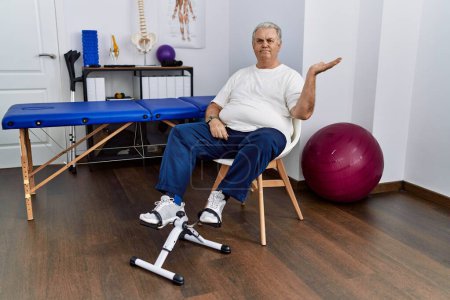 Photo for Senior caucasian man at physiotherapy clinic using pedal exerciser smiling cheerful presenting and pointing with palm of hand looking at the camera. - Royalty Free Image