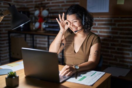 Téléchargez les photos : Young hispanic woman working at the office at night doing ok gesture with hand smiling, eye looking through fingers with happy face. - en image libre de droit