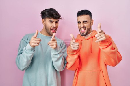Foto de Joven pareja gay hispana de pie sobre fondo rosa señalando los dedos a la cámara con cara alegre y divertida. buena energía y vibraciones. - Imagen libre de derechos