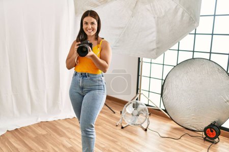 Photo for Young beautiful hispanic woman photographer holding professional camera photo studio - Royalty Free Image
