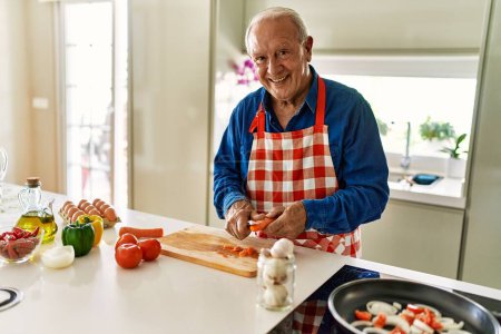 Foto de Hombre mayor sonriendo confiado cortando zanahoria en la cocina - Imagen libre de derechos