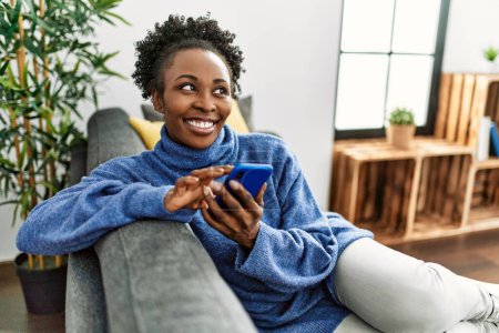 Téléchargez les photos : Femme afro-américaine utilisant un smartphone assis sur le canapé à la maison - en image libre de droit