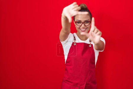 Photo for Young hispanic woman wearing waitress apron over red background smiling making frame with hands and fingers with happy face. creativity and photography concept. - Royalty Free Image