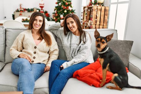 Photo for Two women smiling confident sitting with dog by christmas tree at home - Royalty Free Image