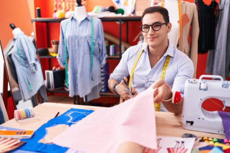 Téléchargez les photos : Jeune homme hispanique tailleur sourire tissu de coupe confiant à l'atelier - en image libre de droit