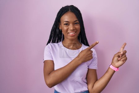 Photo for African american woman with braids standing over pink background smiling and looking at the camera pointing with two hands and fingers to the side. - Royalty Free Image