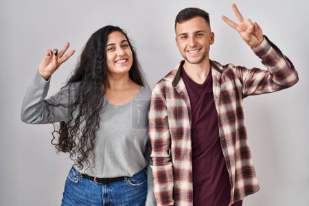Téléchargez les photos : Jeune couple hispanique debout sur fond blanc souriant regardant la caméra montrant des doigts faisant signe de victoire. numéro deux. - en image libre de droit