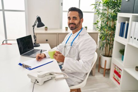 Téléchargez les photos : Jeune homme hispanique portant l'uniforme de médecin écrivant sur le presse-papiers boire du café à la clinique - en image libre de droit
