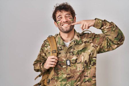Téléchargez les photos : Hispanic young man wearing camouflage army uniform smiling cheerful showing and pointing with fingers teeth and mouth. dental health concept. - en image libre de droit