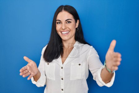 Téléchargez les photos : Jeune femme hispanique debout sur fond bleu regardant la caméra souriant à bras ouverts pour un câlin. expression joyeuse embrassant le bonheur. - en image libre de droit