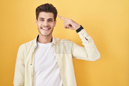 Foto de Joven hombre hispano de pie sobre fondo amarillo sonriendo señalando a la cabeza con un dedo, gran idea o pensamiento, buena memoria - Imagen libre de derechos