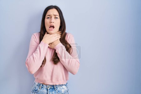 Photo for Young brunette woman standing over blue background shouting and suffocate because painful strangle. health problem. asphyxiate and suicide concept. - Royalty Free Image
