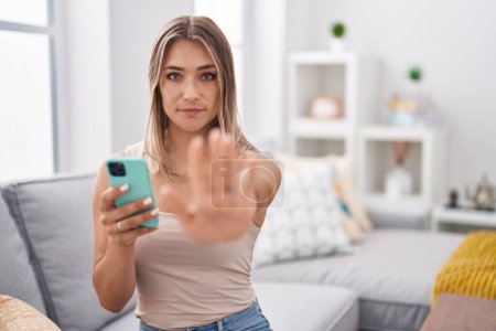 Téléchargez les photos : Blonde caucasian woman using smartphone typing message sitting on the sofa with open hand doing stop sign with serious and confident expression, defense gesture - en image libre de droit