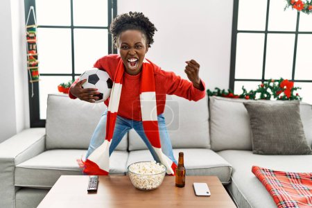 Téléchargez les photos : Femme afro-américaine soutenant l'équipe de football assis sur le canapé à la maison - en image libre de droit