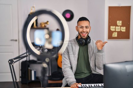 Foto de Young hispanic man playing piano at music studio recording himself pointing thumb up to the side smiling happy with open mouth - Imagen libre de derechos