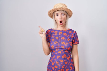 Foto de Mujer joven caucásica con vestido de flores y sombrero de verano sorprendido señalando con el dedo hacia un lado, boca abierta expresión asombrada. - Imagen libre de derechos
