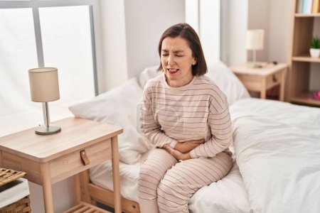 Téléchargez les photos : Femme d'âge moyen souffrant de douleurs menstruelles assise sur le lit dans la chambre - en image libre de droit