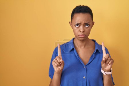 Photo for African american woman standing over yellow background pointing up looking sad and upset, indicating direction with fingers, unhappy and depressed. - Royalty Free Image