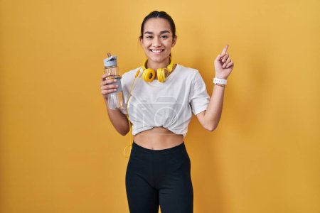 Photo for Young south asian woman wearing sportswear drinking water with a big smile on face, pointing with hand finger to the side looking at the camera. - Royalty Free Image