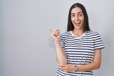 Téléchargez les photos : Jeune femme brune portant un t-shirt rayé avec un grand sourire sur le visage, pointant du doigt vers le côté en regardant la caméra. - en image libre de droit