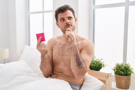 Téléchargez les photos : Jeune homme hispanique tenant le préservatif dans le lit visage sérieux en pensant à la question avec la main sur le menton, réfléchi sur l'idée déroutante - en image libre de droit