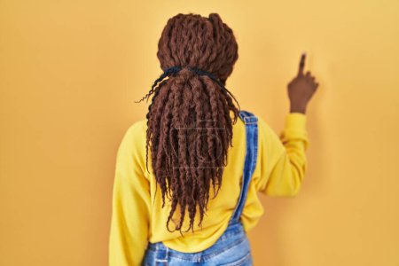 Photo for African woman standing over yellow background posing backwards pointing ahead with finger hand - Royalty Free Image