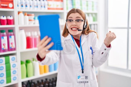Foto de Redhead woman working at pharmacy drugstore doing video call with tablet screaming proud, celebrating victory and success very excited with raised arm - Imagen libre de derechos