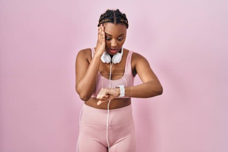 Téléchargez les photos : Femme afro-américaine avec des tresses portant des vêtements de sport et des écouteurs regardant l'heure de la montre inquiète, peur de se retrouver en retard - en image libre de droit