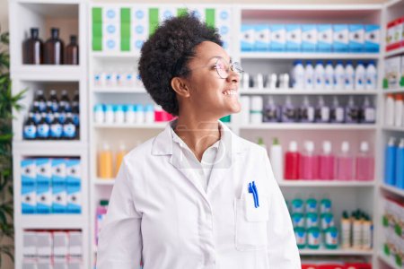Photo for Beautiful african woman with curly hair working at pharmacy drugstore looking away to side with smile on face, natural expression. laughing confident. - Royalty Free Image