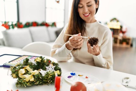Photo for Middle age hispanic woman painting christmas pineapple decor sitting on the table at home - Royalty Free Image