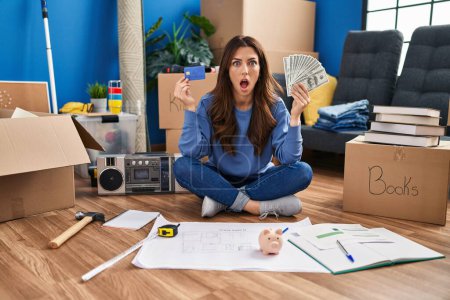 Téléchargez les photos : Young brunette woman sitting on the floor at new home holding savings in shock face, looking skeptical and sarcastic, surprised with open mouth - en image libre de droit
