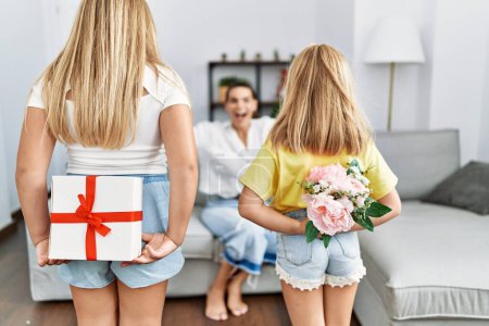 Téléchargez les photos : Mère et filles souriant surprise confiante avec cadeau et fleurs sur le dos à la maison - en image libre de droit