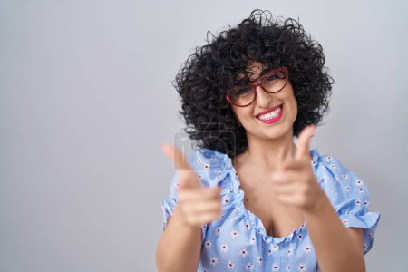 Foto de Young brunette woman with curly hair wearing glasses over isolated background pointing fingers to camera with happy and funny face. good energy and vibes. - Imagen libre de derechos