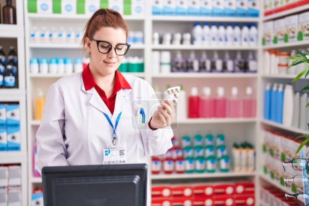 Téléchargez les photos : Young beautiful plus size woman pharmacist using computer holding pills tablet at pharmacy - en image libre de droit