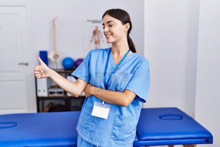 Téléchargez les photos : Jeune femme hispanique portant un uniforme de physiothérapeute debout à la clinique l'air fier, souriant en faisant les pouces vers le haut geste sur le côté - en image libre de droit