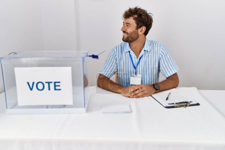 Photo for Young handsome man at political election sitting by ballot looking to side, relax profile pose with natural face with confident smile. - Royalty Free Image