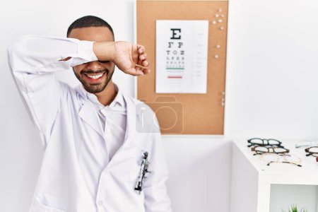 Téléchargez les photos : African american optician man standing by eyesight test covering eyes with arm smiling cheerful and funny. blind concept. - en image libre de droit