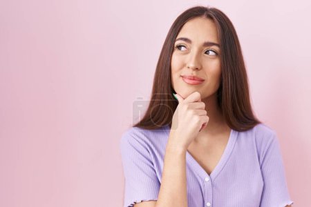 Photo for Young hispanic woman with long hair standing over pink background looking confident at the camera smiling with crossed arms and hand raised on chin. thinking positive. - Royalty Free Image