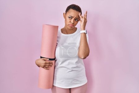Téléchargez les photos : Belle femme afro-américaine tenant tapis de yoga inquiet et stressé par un problème avec la main sur le front, nerveux et anxieux pour la crise - en image libre de droit