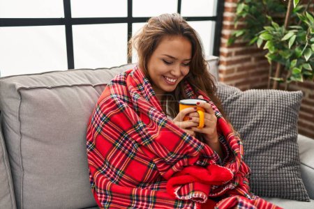 Photo for Young beautiful hispanic woman drinking coffee covering with blanket at home - Royalty Free Image