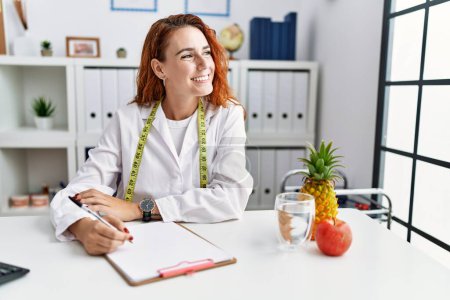 Photo for Young redhead woman nutritionist doctor at the clinic looking away to side with smile on face, natural expression. laughing confident. - Royalty Free Image