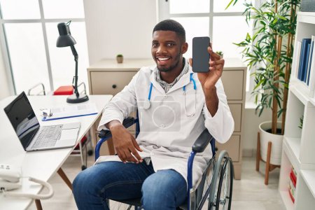 Téléchargez les photos : Afro-américain médecin homme assis sur fauteuil roulant tenant smartphone regardant positif et heureux debout et souriant avec un sourire confiant montrant les dents - en image libre de droit