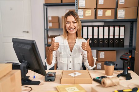 Photo for Young blonde woman working at small business ecommerce success sign doing positive gesture with hand, thumbs up smiling and happy. cheerful expression and winner gesture. - Royalty Free Image