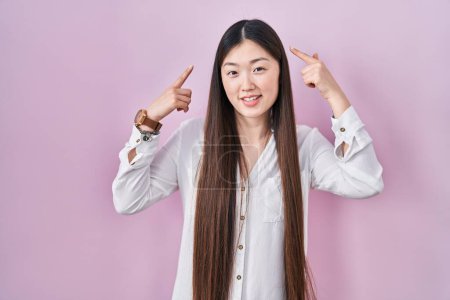 Photo for Chinese young woman standing over pink background smiling pointing to head with both hands finger, great idea or thought, good memory - Royalty Free Image