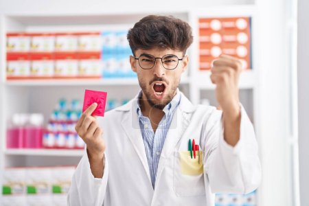 Téléchargez les photos : Homme arabe avec barbe travaillant à la pharmacie pharmacie tenant préservatif peur et choqué avec surprise et expression étonnante, peur et visage excité. - en image libre de droit