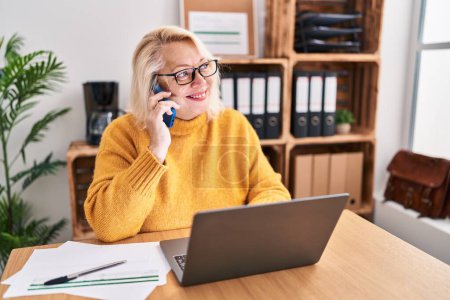 Foto de Mujer rubia de mediana edad mujer de negocios hablando en el teléfono inteligente que trabaja en la oficina - Imagen libre de derechos