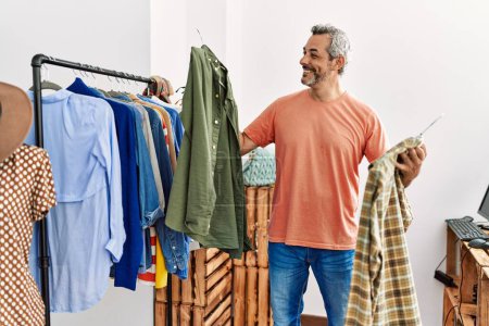 Foto de Hombre de pelo gris de mediana edad cliente sonriendo confiado sosteniendo la ropa de rack en la tienda de ropa - Imagen libre de derechos